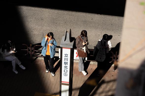 Four people stand on a sunlit sidewalk next to a sign with Japanese text. Each person is engrossed in their phone, wearing casual clothing. Shadows create contrasting light and dark areas. The scene suggests a public waiting area.