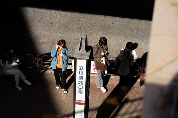 Four people stand on a sunlit sidewalk next to a sign with Japanese text. Each person is engrossed in their phone, wearing casual clothing. Shadows create contrasting light and dark areas. The scene suggests a public waiting area.