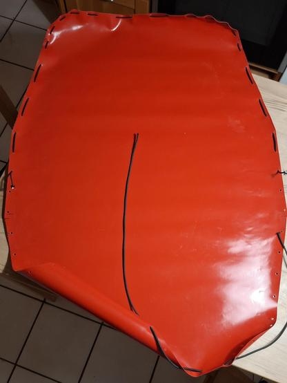 Auf einem Tisch liegt eine zugeschnittene rote Lkw-Plane, die Ränder sind mit Löchern versehen, durch die ein schwarzes Gummiband gefädelt wird.