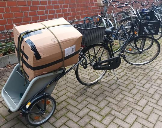 Hinter einem schwarzen Hollandrad mit einem Korb vorne und einem Korb hinten auf den Gepäckträgern ist ein 35 Jahre alter Donkey-Einkaufsfahrradanhänger angekoppelt auf dem ein großes Paket festgeschnallt ist, dass das Ladevolumen des Donkeys bei weitem übersteigt. Im Hintergrund eine Fahrradabstellanlage mit abgestellten Fahrrädern vor einer Backsteinmauer.