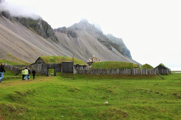 View on a small viking village, formed from some wooden huts, surrounded by wooden palisades. On the right in the background is a mountain slope.