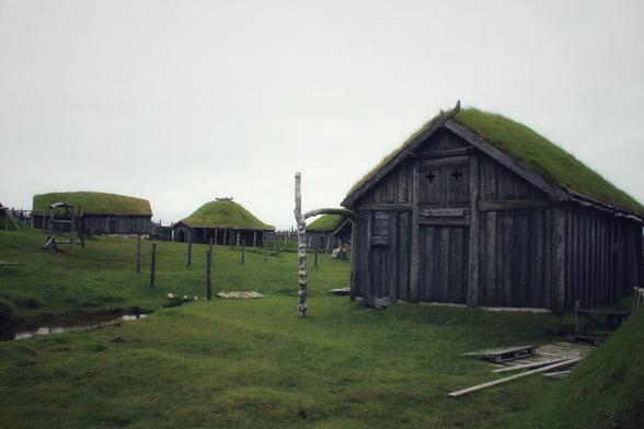 Some wooded huts with green overgrown roofs. One hut is on the right in ther foreground two are in the back. the huts stand on a green meadow.