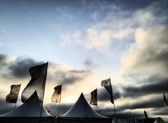 In der unteren Hälfte des Bildes ragen zwei spitze weiße Zeltdächer und dahinter fünf weiße, rechteckige im Wind wehende Flaggen in den bewölkten Abendhimmel. Im Gegenlicht der untergehenden Sonne im Hintergrund wirken diese dunkel bis schwarz- silhouettenhaft vor den ebenfalls dunklen Wolken im unteren Bereich des Himmels. Darüber sind die Wolken noch weiß und der Himmel hellblau. 

In the lower half of the picture, two pointed white tent roofs and, behind them, five white rectangular flags blowing in the wind rise into the cloudy evening sky. Backlit by the setting sun in the background, these appear dark to black, silhouetted against the equally dark clouds in the lower part of the sky. Above them, the clouds are still white and the sky is light blue.