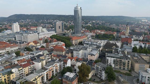 Dieses Luftbild zeigt eine Stadtlandschaft. Es gibt eine Vielzahl von Gebäuden unterschiedlicher Höhe und Architektur, darunter Wohnhäuser, Bürogebäude und ein hoher Turm in der Mitte des Bildes. Der Turm ist silbrig und hat eine vertikale Streifenstruktur. Im Hintergrund ist ein großer, grüner Hügel zu sehen. Die Gebäude sind unterschiedlich farbig, mit viel Rot und Weiß, und stehen dicht aneinander. Die Sonne scheint auf die Stadtlandschaft.