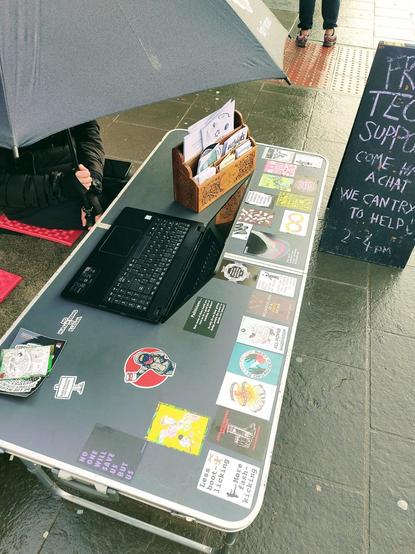 A person, face obscured by a large umbrella, sits in front of a laptop on a pop-up outdoor table. The table is adorned with colourful anarchist stickers.

In the background there is a black pop-up board with chalk writing, text partially obscured by the edge of the image. It reads:

"Free Tech Support. Come have a chat. We can try to help 🙂. 2-4PM"