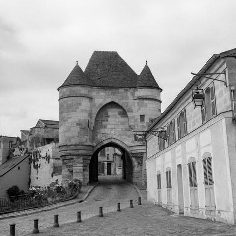 Black and white photo of a large medieval stone gate. A winding road leads through the gate into a city with historic buildings.