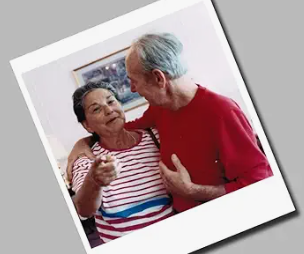 my mom wearing a red and white striped t-shirt pointing at the camera. My dad looking at her with his arm around her. He is wearing a red shirt. Not looking at the camera.