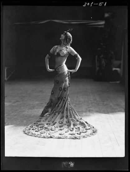 This black and white photograph captures a woman performing what appears to be an exotic dance, likely inspired by Middle Eastern or Indian styles. She is dressed in a shimmering costume that features sequins, beads, and intricate patterns on her bodice, waistband, and headpiece. The skirt of the dress extends into an elaborate train with layers resembling peacock feathers, which adds drama to the performance. Her hands are placed firmly on her hips as she leans back slightly, displaying a pose filled with grace and elegance.

The background is nondescript, possibly indicating that this photo was taken in a studio or at a rehearsal space rather than during an actual performance. The focus is solely on the dancer's costume and posture, emphasizing the artistry of both the garment and her dance technique. There are no other individuals visible in the image to distract from the subject.

The photograph carries historical significance as it provides insight into early 20th-century fashion, stage costumes, and possibly cultural performances during that era. The name "St. Denis" could refer to an individual or a group associated with this performance, while "Ruth Gente..." is partially visible but not clear enough for further interpretation.

This image would be suitable as alt text because it captures the essence of the subject matter without relying on color, which often cannot accurately convey such visual information in mo [...]