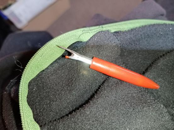 A sewing picker in red on top of the green zipper that is partially unpicked.
