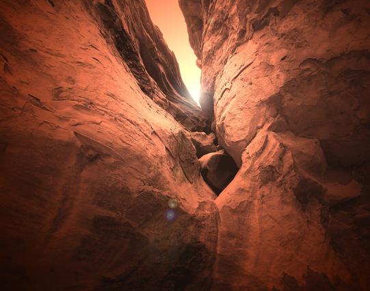 A color photo showing a low angle view of a break in a rock formation. The scene is bathed in sunlight that is peeking through the break in the rocks.