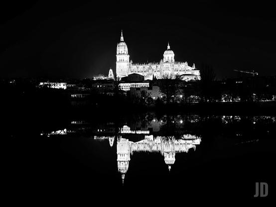 Fotografía nocturna en blanco y negro que captura un impresionante paisaje urbano con un edificio monumental reflejado en el agua.
• Sujeto Principal: El elemento central es una gran catedral o iglesia majestuosamente iluminada, que se alza sobre el horizonte de la ciudad. Se distinguen claramente sus torres, cúpulas y la arquitectura detallada, contrastando fuertemente con la oscuridad del cielo nocturno.
• Composición: La parte inferior de la foto muestra un reflejo casi perfecto y muy nítido del edificio en la superficie de un cuerpo de agua (posiblemente un río o lago). Este reflejo crea una poderosa simetría.
• Estilo y Tono: El uso del blanco y negro intensifica el drama y la solemnidad de la escena. La iluminación de la estructura le da un aire etéreo, casi flotante, mientras que el resto del primer plano y el fondo están dominados por la oscuridad (siluetas de árboles y tejados).
• Identificación: Por la arquitectura y el reflejo, se identifica claramente como la Catedral de Salamanca (España), vista desde el otro lado del río Tormes.