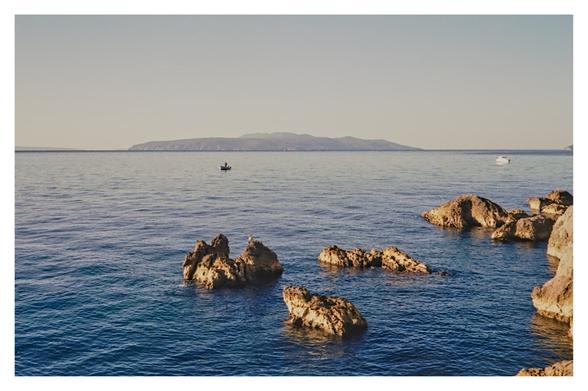Color photograph of the Adriatic Sea with a view of the island of Cres, taken from a bathing spot between Ika and Ičići. Although taken in the morning, the light is already very bright. The small rocks in the foreground glow bright gold, and the sky ranges from turquois to almost white on the horizon. Two small boats float in the water.