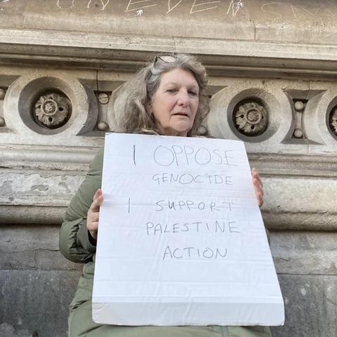 Woman with greying hair sitting on the ground outside the Royal Courts Of Justice in London, holding a large handwritten sign "I oppose genocide. I support Palestine Action".