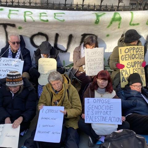 A group of 8 people, mostly women, sitting on the ground outside the Royal Courts Of Justice in London, holding a large handwritten sign "I oppose genocide. I support Palestine Action".