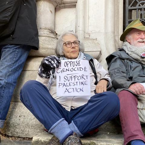 Woman with grey hair sitting on the ground at the foot of a pillar outside the Royal Courts Of Justice in London, holding a large handwritten sign "I oppose genocide. I support Palestine Action".  On her left side sits an elderly man.