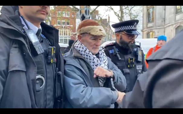 Outside the Royal Courts Of Justice in London, an elderly man in handcuffs is led away by two uniformed police officers