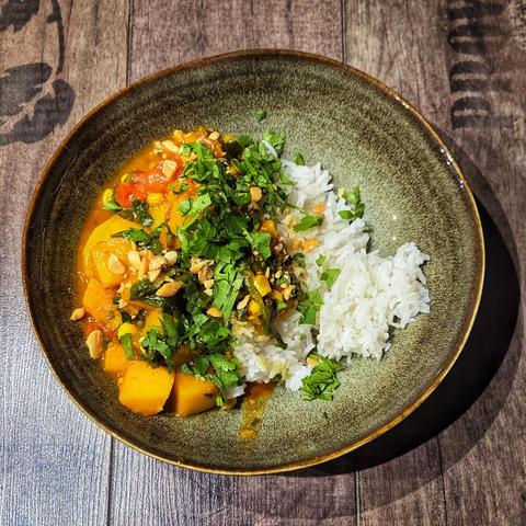 Ein gelbliches Curry Gericht mit Reis in einer Terrakotta Schale, bestreut mit gehackten Erdnüssen und Koriandergrün