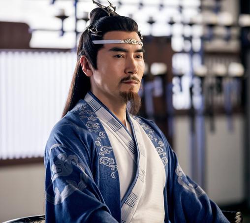 Lan Qiren sits with impeccable posture and equally impeccable goatee, hands resting on his needs just out of frame below the bottom edge of the image, and his gaze focused on a target beyond the viewer. His loose overrobe is a rich blue embroidered with the Gusu Lan sect symbol on the upper arms of the wide sleeves and trimmed with cloud shaped embroidery from mid-chest on up around the collar. The outer robe layered underneath the overrobe is white with a thin blue edge abutted by a wider strip featuring regular spaced rectangles composed of blue dots. A thin sliver of white from an underlayer can be seen at the throat. He wears the customary sacred Lan forehead ribbon complete with inner disciple exclusive emblematic metal piece in the middle. The crown attached to his hairbun has twin forks, while another hairpiece complementary in design wraps around the back of his head below the bun, reaching to just behind his temple on either side.