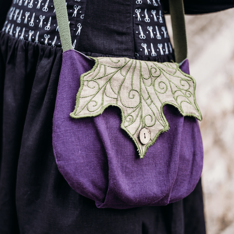 Gros plan sur un sac en bandoulière en forme de grosse aubergine ronde. Le rabat est fait en lin vert avec des nervures brodées, le corps est en lin violet et la hanse est en coton vert.
