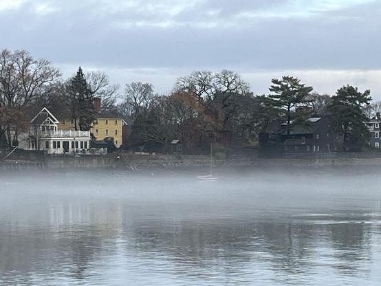 Fog hangs over Salem Harbor. Behind the fog is a sea wall. Above the sea wall are three noticeable houses. To the left is a large yellow house and a blue house with a white pillared porch. To the right, slightly obscured by trees is a dark brown/black low two-story house that is the House of Seven Gables.