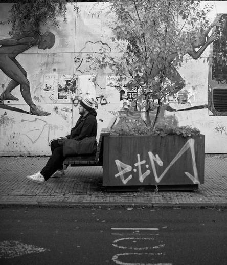 A person sits on a bench in front of a graffiti-covered wall featuring a large mural of a figure in motion. Nearby, a planter with greenery adds a touch of nature to the urban scene. The image is in black and white.