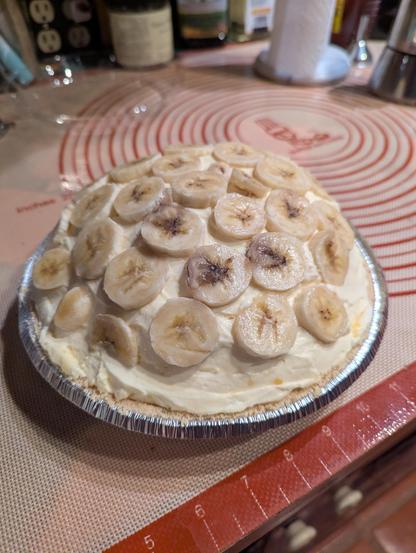 A 9" pie with a graham cracker crust filled with folded vanilla pudding and fresh whipping cream. The cream is mounded super high and topped with fresh banana slices.