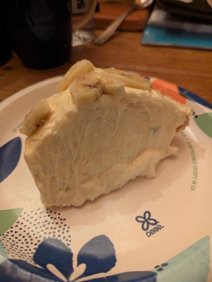 A piece of banana cream pie atop a Dixie paper plate. The pie is tall and creamy with fresh banana slices on top.