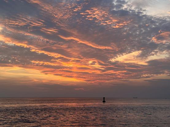 A serene sunset over the ocean, featuring vibrant hues of orange and pink in the sky with scattered clouds. A buoy is visible in the foreground, and distant ships can be seen on the horizon.