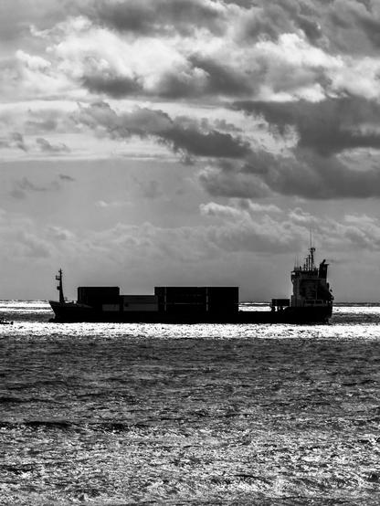 Auto-generated description: A cargo ship sails across a vast ocean under a dramatic, cloudy sky.