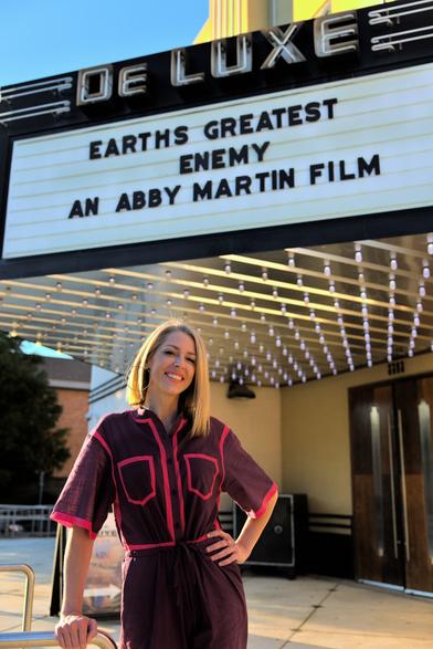 Abby Martin standing outside of the theater with the backdrop above her stating "Earths Greatest Enemy - An Abby Martin Film"