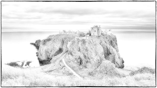 A high key black and white image of a rocky coastal landscape featuring the ruins of a castle on a cliff overlooking a calm sea, with a winding path leading toward the structure. The sky appears cloudy and bright, creating a serene atmosphere.