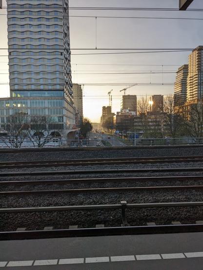 Standing on a train platform, a street goes underneath, tall office buildings line the road, some cranes in the distance still working on some. Meanwhile the sun is about to set, but her golden rays still blanket everything in gold