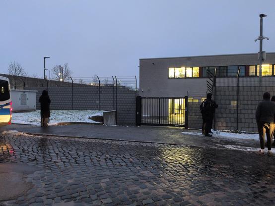 Links im Bild ist ein kleiner Teil eines weiß-blauen VW-Bus der der Polizei zu sehen. Im.Vordergrund gepflasterte Strasse, dahinter ein Tor, Gitter und ein zweistöckiger Flachbau mit beuchteten Fenstern. Es ist ein Bild in der Abenddämmerung

Zu sehen sind zwei maskierte Polizisten und drei andere Menschen. Dazu noch etwas Schnee.

Das Bild ist vor dem OLG Dresden, Aussenstelle Hammerweg, aufgenommen. Die Menschen die dort stehen, warten auf den Abtransport der Angeklagten im Fall 'Antifa-Ost', die von massiven Polizeikräften in diverse Gefängnisse eskortiert werden.