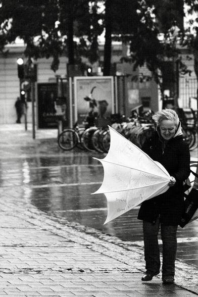 Das Foto zeigt eine Straßenszene in Schwarz-Weiß in der St. Pöltenter Innenstadt an einen regnerischen windigen Tag. Man sieht eine Person mit Regenschirm der durch den Wind verbogen ist.