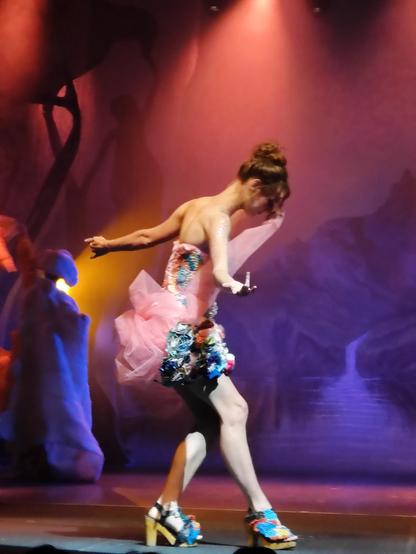 Woman clothed in a short strapless blue dress adorned with pink tulle, arms outstretched dancing across the stage.