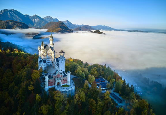 An aerial view of Bavaria’s Neuschwanstein Castle perched above an autumn-colored forest, with a sea of clouds rolling over the valley below