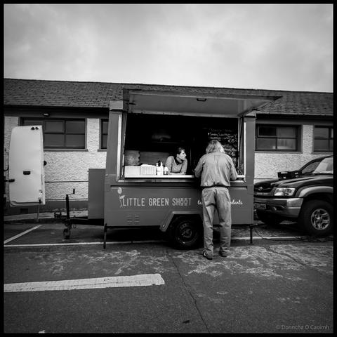 Black and white photograph of a food trailer called "Little Green Shoot" at Skibbereen Market with a customer standing at the service window being served by staff inside, a menu board displaying prices including "€5", surrounded by a parked car on the right and a van door visible on the left, and a single-storey building under overcast sky.