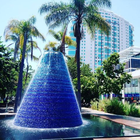 a water volcano is seen with some palm trees and some buildings behind