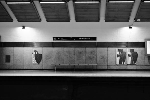 BW picture of an empty bench at the Martim Moniz Subway station. Above the bench a sign identifying the Station by its name.