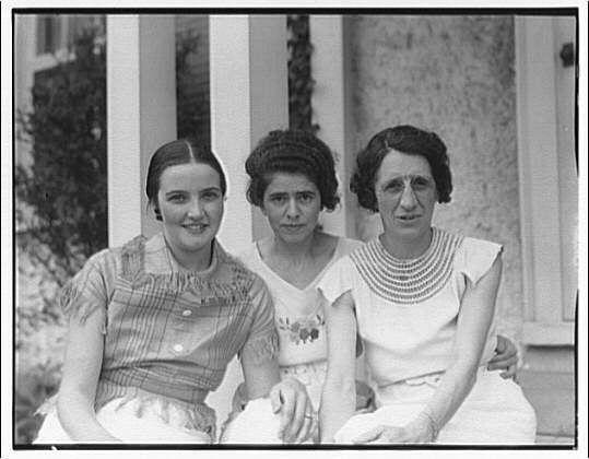 The image is a black and white photograph showing three women sitting closely together on what appears to be a porch. The woman in the middle has her hair styled with bangs, pulled back into an updo, wearing a patterned dress with short sleeves. She sits beside another woman who also wears a dress featuring decorative elements such as ruffles or lace at the neckline and hem; she is smiling gently.
To the right of this pair is a third woman sitting in a more reserved pose, dressed in a white outfit adorned with a prominent beaded necklace that reaches down to her mid-chest. This woman's expression is neutral but appears slightly serious compared to the others' smiles or relaxed demeanor.
The background reveals part of what seems like an exterior wall made from textured material and some vertical supports which might indicate pillars, suggesting a residential setting. The photograph has been noted as belonging in the collection of Eveline Horydczak (1890-1971), with additional context provided by "Eveline" referenced below.
The image is credited to Horydczak himself and indicates that it was likely taken around a period between 1920 and 1950. The photograph depicts the women, their attire, and overall presentation reflecting fashion trends from early-to-mid twentieth-century America.