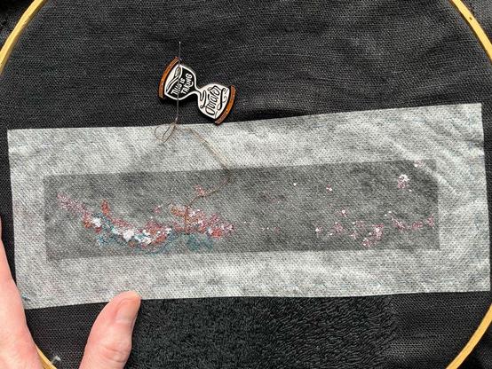 Lia’s fingers show a wooden embroidery hoop with black linen with a rectangle of dissolvable material on which is printed the Milky Way in black and white with some red and blue markings. Some tiny single strand seed stitch and French knots in white, peach, oranges, and dark browns and grays have been stitched. Below the Milky Way is black on black whipped backstitch that looks a bit like flames, it’s pubic hair. An hourglass-shaped needle minder that reads “this is taking forever” is attached to the cloth.