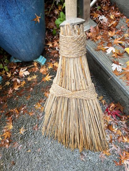 Closer view of the broom. It has a triangular head made from split stiff plant material not grass but something split from cane or wood. It's held together with woven twine and a ferrule around the hardwood handle made of coconut shell.
