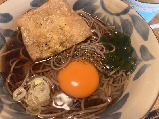 A bowl of soba noodles in a dark broth, topped with a raw egg, a piece of fried tofu, seaweed, and sliced green onions. Various seasonings are sprinkled on top.