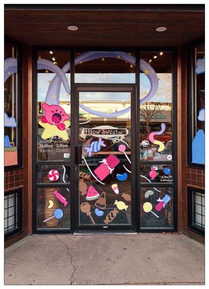 Double glass doors of a candy shop adorned with colorful cutouts of candies and sweets, including lollipops, ice cream cones, and cupcakes. The doors feature a purple banner with yellow stars across the top and are framed by dark wooden panels. The shop's address, 314, and hours are displayed on the doors. Reflective glass shows a bit of the street and other buildings outside.