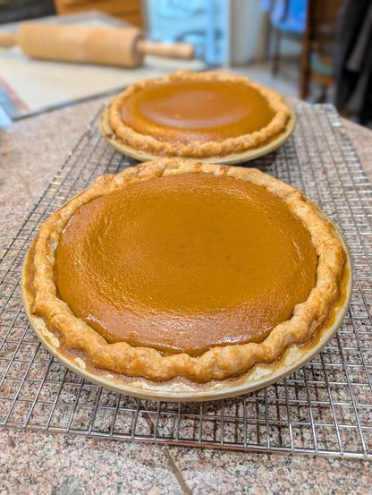 Brown homemade pumpkin pie, fresh from the oven, on a cooling rack. A second pie is in the background.