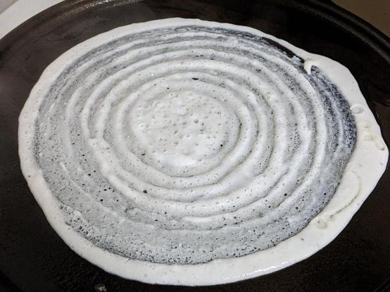 Dosai batter spread out in a spiralized circle on a dark cast iron griddle