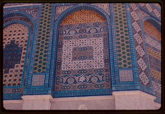 The image displays a segment of an intricate mosaic pattern, likely part of the historic Dome of the Rock in Jerusalem. The design is rich with geometric and floral motifs set against a background that combines deep blue, white, gold, and hints of green tiles. These patterns are framed by bands of alternating colors, creating a rhythmic visual flow across the surface.

Several arched windows or openings can be seen, which allow light to filter into the space they occupy. The craftsmanship suggests an Islamic architectural influence, characterized by its elaborate tilework and use of Arabic calligraphy within some sections. This particular photograph appears aged with a yellowish tint indicative of vintage film photography from around 1950-1977.

There is no visible human activity or modern elements in the frame, emphasizing the timeless beauty and historical significance of this architectural marvel.
