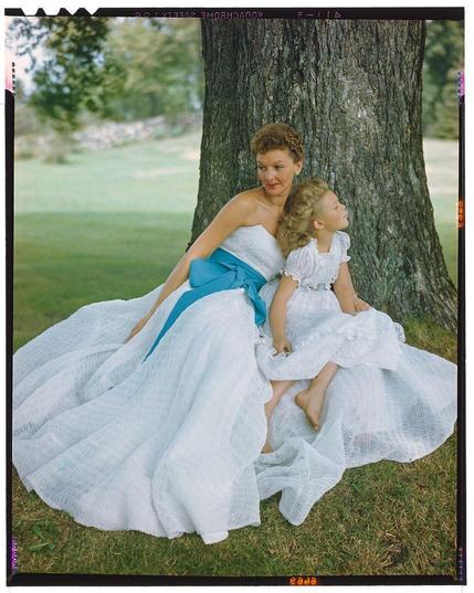 A color photograph of two women dressed in white dresses sitting under a tree, with the woman on the left wearing a blue sash. The photo was taken by Gjon Mili for Life magazine and is dated August 1949; it appears to be a scene depicting Mary Martin spending time outdoors with her daughter Heller Frissell.