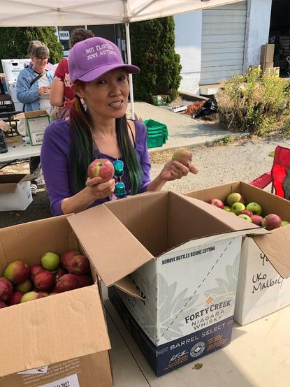 Me, with rainbow-colored hair and a purple hat that says "NOT FLIRTING JUST AUTISTIC" holds two apples in front of several cardboard boxes of apples, outside of LifeCycles office.