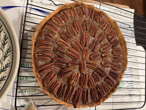 A freshly baked pecan pie topped with a decorative arrangement of whole pecans. The pie sits on a wire cooling rack, with a patterned plate visible in the background.